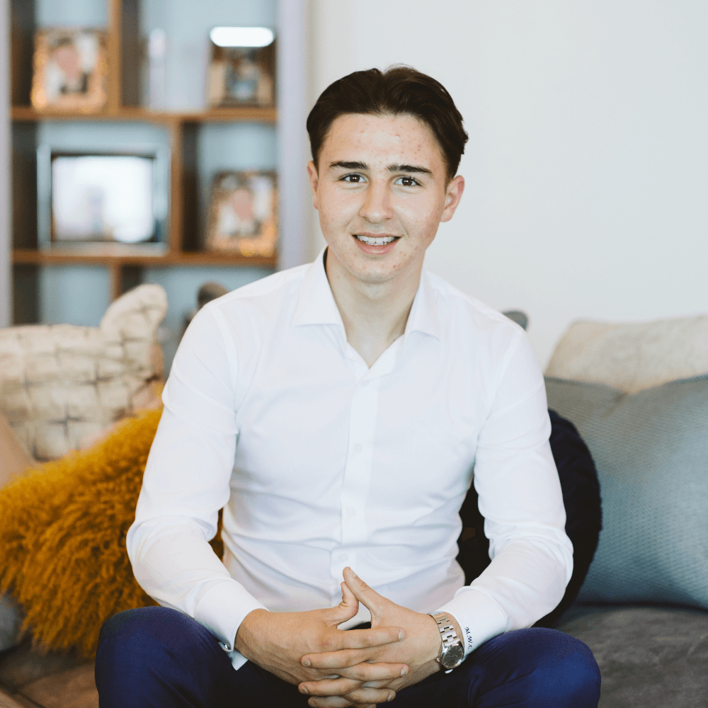 Smiling young man in a white dress shirt sitting on a sofa with hands clasped.